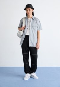 Lys grå kortærmet skjorte, sorte bukser med stikninger, hvide sneakers og en sort bucket hat. Skuldertaske i lys grå.