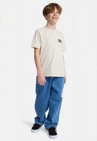 T-shirt crème à manches courtes avec un petit logo noir et blanc sur la poitrine, associée à un jean en denim bleu ample et des baskets noires.