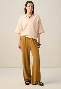 T-shirt oversized rose clair à col en V associé à un pantalon large moutarde. Tissu texturé et design décontracté. Sandales plates.
