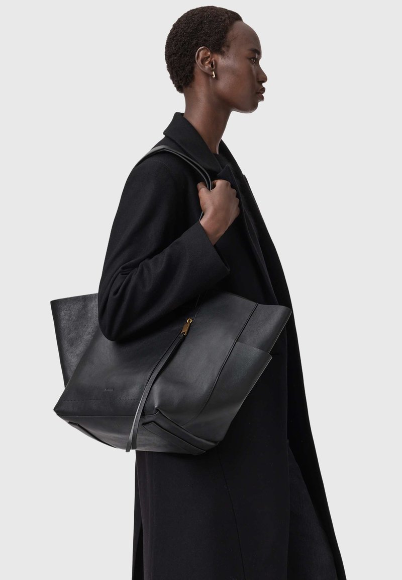 Sac fourre-tout en cuir noir avec un design angulaire, une texture lisse et un détail de fermeture éclair subtil. Porté à l'épaule, complétant un manteau noir.