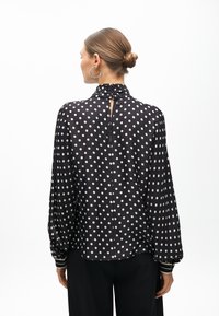 Schwarze Bluse mit weißen Punkten, hoher Kragen, lange Ärmel und Knopfschluss auf der Rückseite. Mit gestreiften Ärmelbündchen. Glatte Stofftextur.