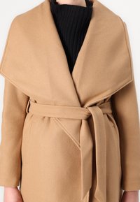 Manteau beige à enrouler avec un large col, taille cintrée et poches latérales. Texture douce et design minimal sans fermetures visibles.