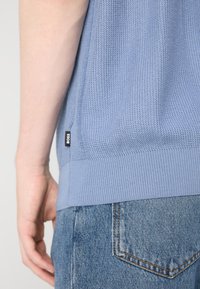 Gros plan sur une personne portant un pull tricoté bleu clair texturé avec une étiquette "BOSS" et un jean en denim bleu avec une poche arrière visible.
