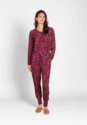 Woman standing wearing a long-sleeve, burgundy floral jumpsuit with drawstring waist and tan slip-on shoes with purple straps.