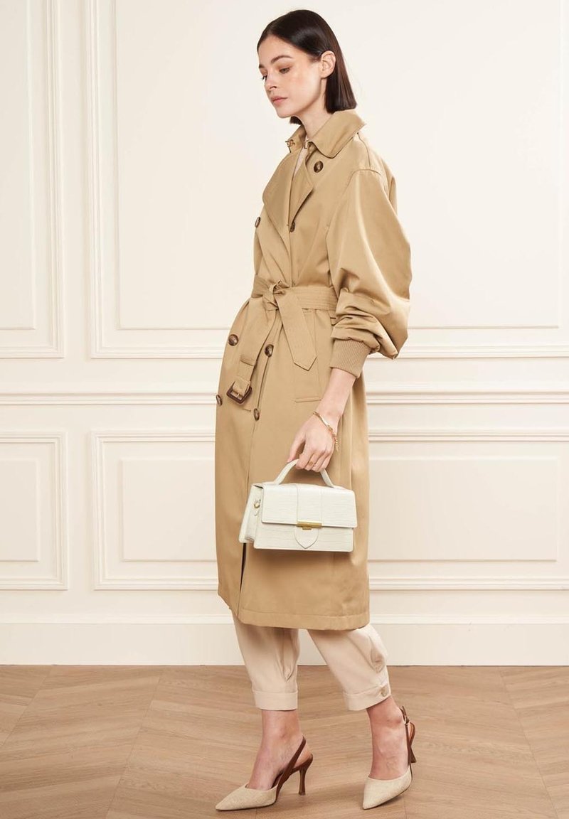 Tan trenchcoat met oversized mouwen, ceintuur in de taille en knoopdetails. Gepaarde met een crème-handtas en beige puntige hakken. Glad, zorgvuldig passend.