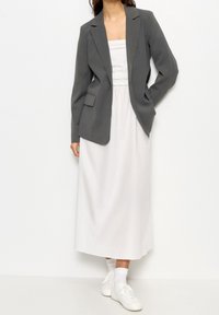 Blazer structuré gris sur une robe maxi blanche. Le blazer présente un col cranté et des poches latérales. Des baskets blanches complètent la tenue.