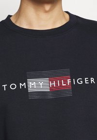 Felpa girocollo blu navy con una grafica di strisce alternate rosse e bianche, con la scritta "TOMMY HILFIGER" stampata in bianco sottostante.