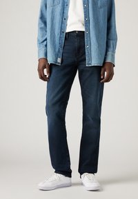 Jean bleu foncé à coupe droite, associé à une chemise en denim clair et des baskets blanches. Tissu lisse avec des détails de couture subtils.