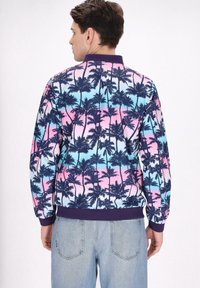 Jachetă bomber colorată, cu un model de palmier în nuanțe de albastru marin pe un fundal degradat de roz și albastru deschis. Manșete și tiv din material elastic.