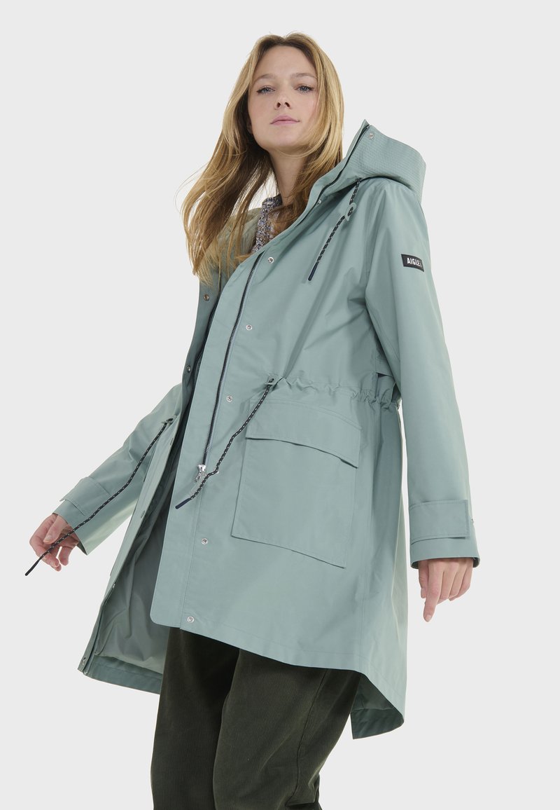 Veste imperméable vert clair avec capuche, taille élastique, poches avant et cordons de serrage réglables. Texture lisse et détails à boutons-pression.