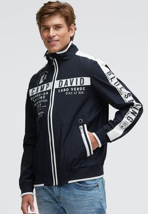 Young man smiling wearing a navy blue sailing jacket with white text and stripes, standing with one hand in pocket against plain background.