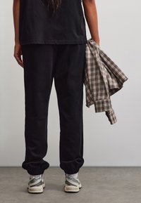 Persona con pantaloni neri e sneakers, che tiene in mano destra una camicia beige e nera a quadri, in piedi su un pavimento grigio.