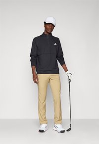 Čierny zipsový pulóver, béžové nohavice a biele golfové topánky. Na hlave má bielu čiapku a drží golfovú palicu. Pulóver má na sebe logo Adidas.