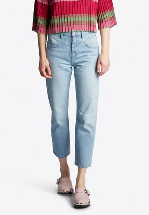 Femme portant un jean court taille haute bleu clair, des chaussures à enfiler roses cloutées, et un pull court en tricot rayé rouge et vert.