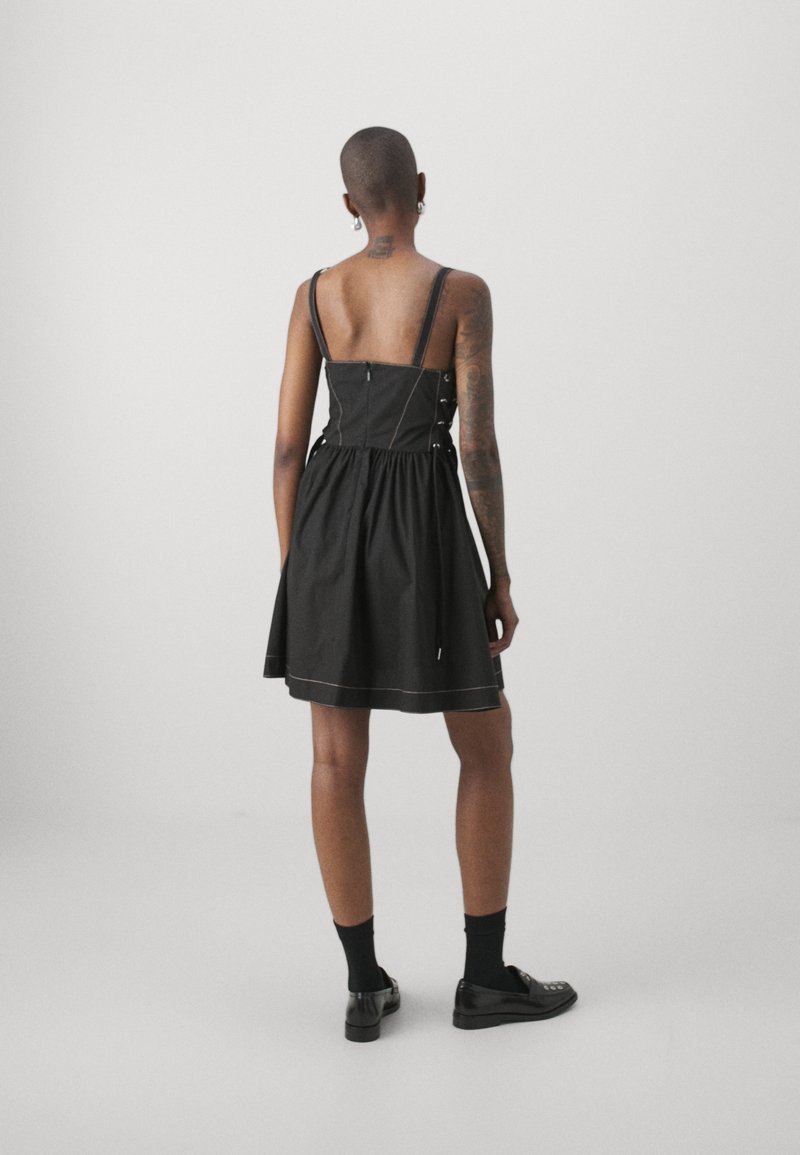 Personne aux cheveux courts et tatouages portant une robe noire sans manches, des chaussettes noires et des chaussures noires, debout, de dos, sur un fond uni.