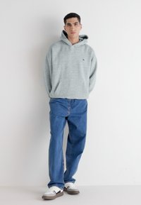 Sudadera gris con capucha y bolsillo frontal, combinada con pantalones vaqueros azules y zapatillas blancas con acentos grises. De pie frente a un fondo sencillo.