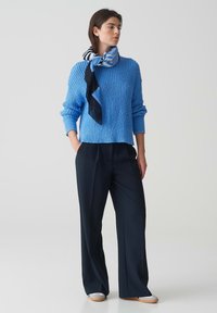 Maglione azzurro chiaro con trama, maniche lunghe, abbinato a pantaloni ampi blu navy. Una sciarpa a fantasia completa il look attorno al collo.