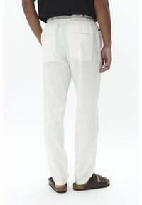 Baltos, lengvos medžiagos kelnės su elastinga juosmens juosta, turinčios du užpakalinius kišenes. Dėvimos su tamsiomis sandalais, demonstruojantys laisvą siluetą.