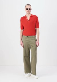Rød strukturert polo shirt med korte ermer, olivengrønne løse bukser og hvite sneakers. Modellen har også runde solbriller.