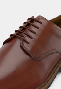 Brown polished leather dress shoe with round laces, detailed stitching, and a smooth sole edge, photographed against a neutral background.