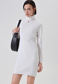 Donna che indossa un vestito aderente bianco a collo alto, grandi orecchini a cerchio, collana con ciondolo d'oro e tiene in mano una borsa a tracolla nera.