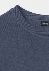 Gros plan du col d’un pull en maille côtelée bleu marine avec une étiquette noire rectangulaire de la marque MNG à l’intérieur du col.