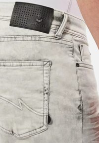 Jean en denim gris délavé avec une étiquette en cuir noir texturé à la taille et des coutures visibles. Comprend une poche arrière avec un design en zigzag.