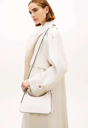 Femme aux cheveux courts et bruns portant un trench blanc, tenant un petit sac à main blanc avec une bandoulière noire sur l'épaule.