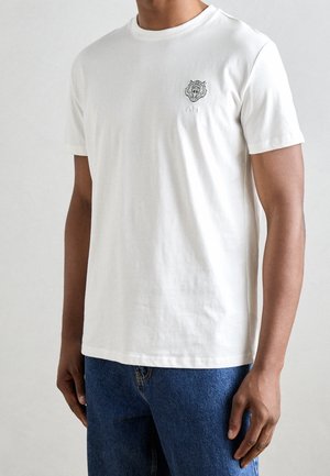 Homme portant un t-shirt blanc uni à manches courtes avec un petit logo tigre sur la poitrine gauche et un jean bleu sur un fond neutre.