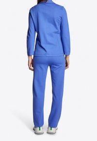 Eine Frau trägt eine passende blaue Jacke und Hose mit weißen Turnschuhen, steht mit dem Rücken zu einer schlichten weißen Hintergrund.