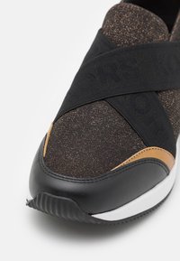 Gros plan sur l'avant d'une chaussure de sport noire et marron avec un tissu scintillant, des sangles élastiques noires en croix, et des détails en caoutchouc blanc et noir sur la semelle.