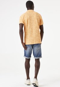 Camisa polo de manga corta en naranja claro con un estampado de hojas, combinada con pantalones cortos de denim enrollados y zapatillas blancas. Vista trasera.