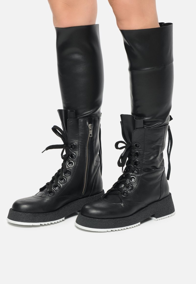 Bottes noires en cuir jusqu'au genou avec une semelle en plateforme texturée, un laçage à l'avant et une fermeture éclair sur le côté. Elles présentent des œillets renforcés et une finition lisse.