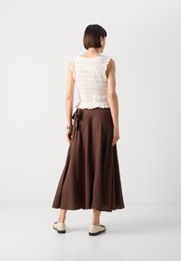 Top bianco smockato con bordi arricciati abbinato a una gonna maxi marrone con un dettaglio in vita annodato. Modella con scarpe chiare.
