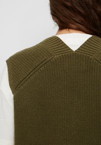 Dziergany bezrękawnik w kolorze oliwkowej zieleni z prążkowaną fakturą. Posiada dekolt w kształcie litery V oraz kontrastujące wzory dzianiny na ramionach.