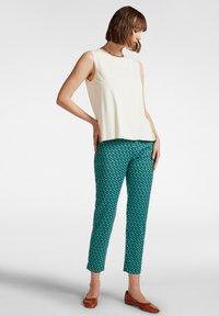 Top bianco senza maniche con pieghe abbinato a pantaloni teal con motivo a ripetizione. Scarpe basse marroni completano l'outfit.