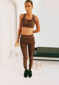 Luipaardprint sportbeha en leggings in bruin en zwart, gemaakt van rekbare stof. Model houdt een doorzichtige waterfles vast; zwarte sneakers aan de voeten.