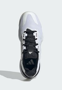 Zapatilla deportiva blanca con acentos negros, parte superior de malla y cordones negros. Cuenta con una lengüeta acolchada y un logotipo en la plantilla. Suela de goma redondeada.