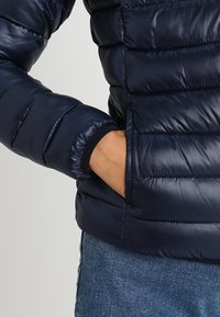 Námořnická pufovaná bunda s prošívanými sekcemi, elastickými manžetami a bo�ční kapsou, vyrobená z hladkého, lesklého materiálu.