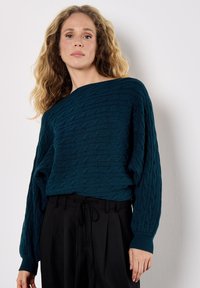 Maglione lavorato a maglia color teal con un motivo a trecce testurizzato, scollatura a barca ampia e maniche lunghe. Abbinato a pantaloni neri a vita alta.