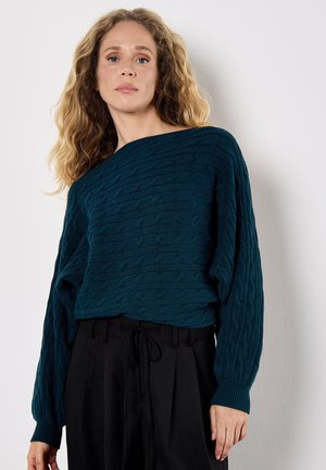 Pull en maille bleu-vert avec un motif de câble texturé, décolleté large bateau et manches longues. Associé à un pantalon noir taille haute.