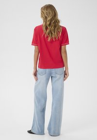 Chemise rouge à manches courtes avec des bordures blanches, associée à un jean large bleu clair. Tissu lisse, coupe décontractée et détails de couture à l'arrière visibles.