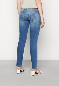 Högmidjade blå denimjeans med smal passform, med dekorativa bakfickor och subtila slitningar; stylade med vita blockklackssandaler.