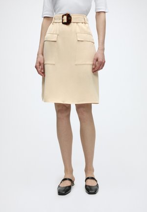 Žena má na sebe béžovú sukňu po kolená s veľkými prednými vreckami a opaskom s prackou v motíve korytnačej škrupiny, v kombinácii s čiernymi balerínami Mary Jane.