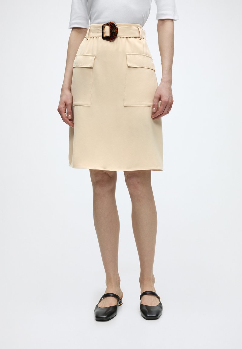 Γυναίκα που φοράει μπεζ φούστα μέχρι το γόνατο με μεγάλες μπροστινές τσέπες και ζώνη με αγκράφα σε σχέδιο χελώνας, σε συνδυασμό με μαύρα παπούτσια Mary Jane με φλατ σόλα.