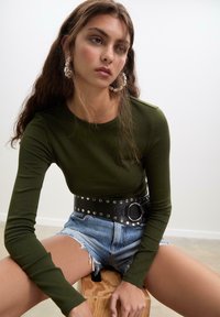 Olivgrünes, eng anliegendes Langarmshirt, kombiniert mit zerrissenen Jeansshorts; schwarzer Gürtel mit großer runder Schnalle und silbernen Nieten, glänzende Creolen.