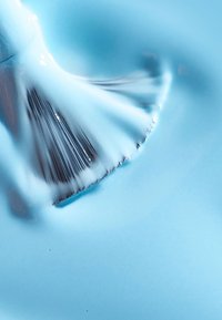 Niebieska farba w płynie o gładkiej, błyszczącej teksturze, rozprowadzana płaskim pędzlem, odsłaniająca cienkie włosie i lśniącą powierzchnię.