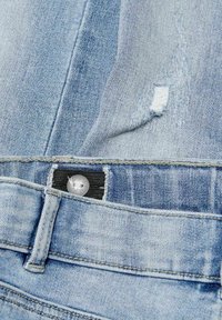 Los jeans de denim azul claro presentan una textura desgastada en la cintura, con un botón plateado y una etiqueta de tela negra cosida en la cintura.