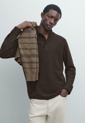 Brun langærmet poloshirt med krave; kombineret med en mønstret beige sweater, der er lagt over skulderen; lyse bukser.