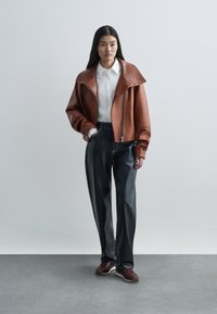 Žena u smeškastom kožnom kaputu preko bele majice, s crnim kožnim hlačama i burgundijskim patikama, stoji uz ravnu pozadinu.